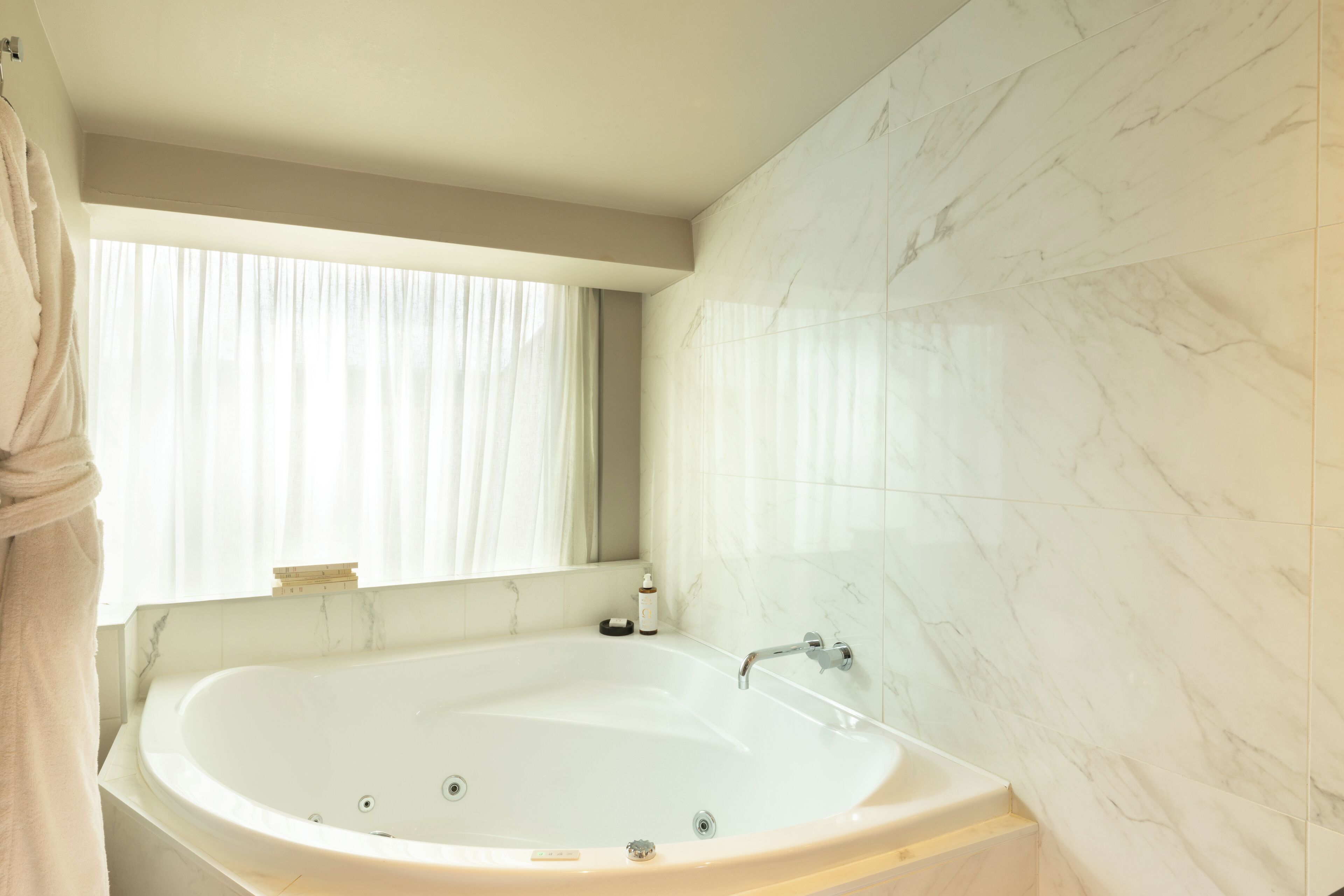 A modern bathroom with a large bathtub, a window with curtains, and marble walls.
