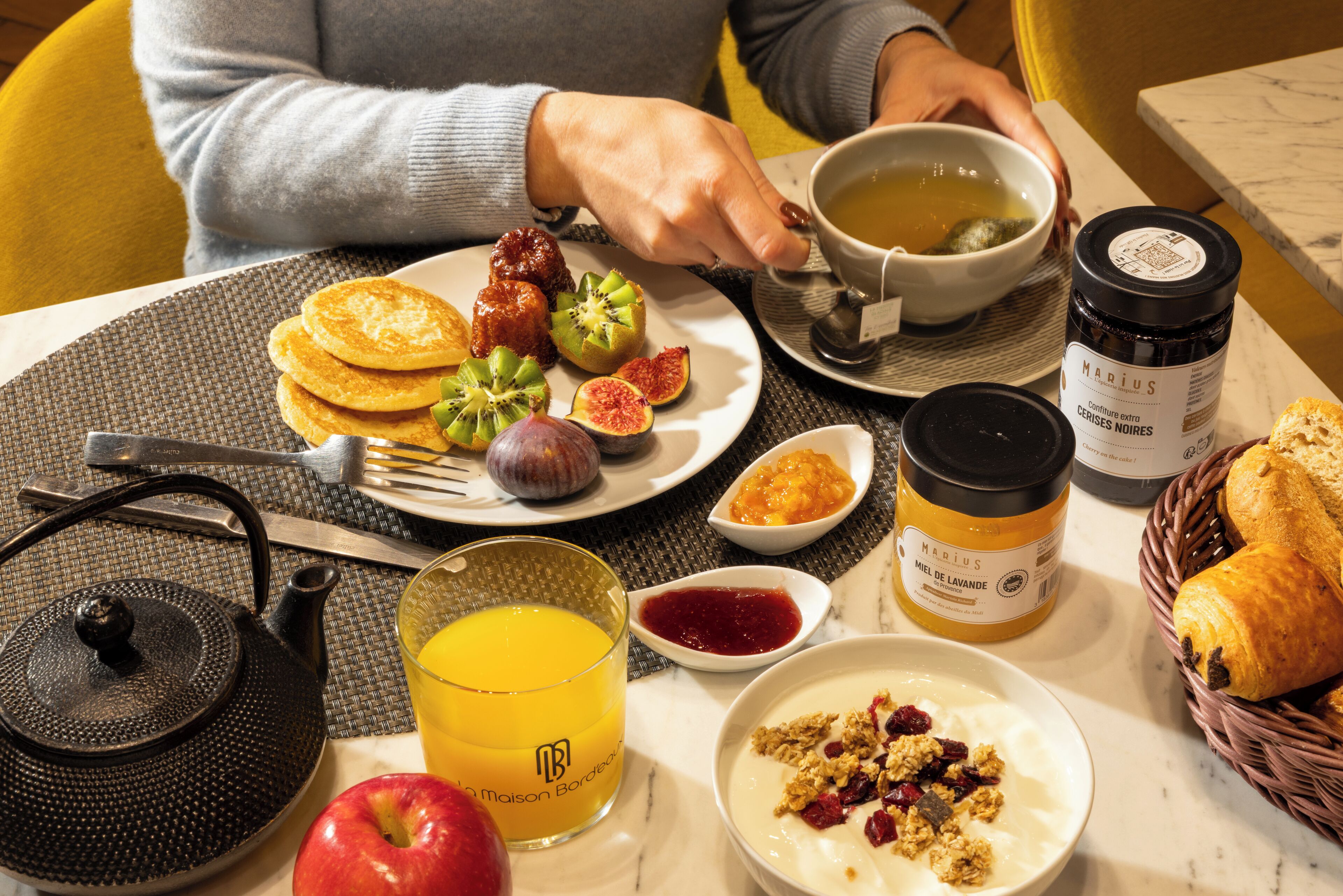 A breakfast setting with a plate of pancakes, fruits, a bowl of soup, a glass of orange juice, a teapot, a jar of honey, a bowl of yogurt with fruit, and a basket of croissants.