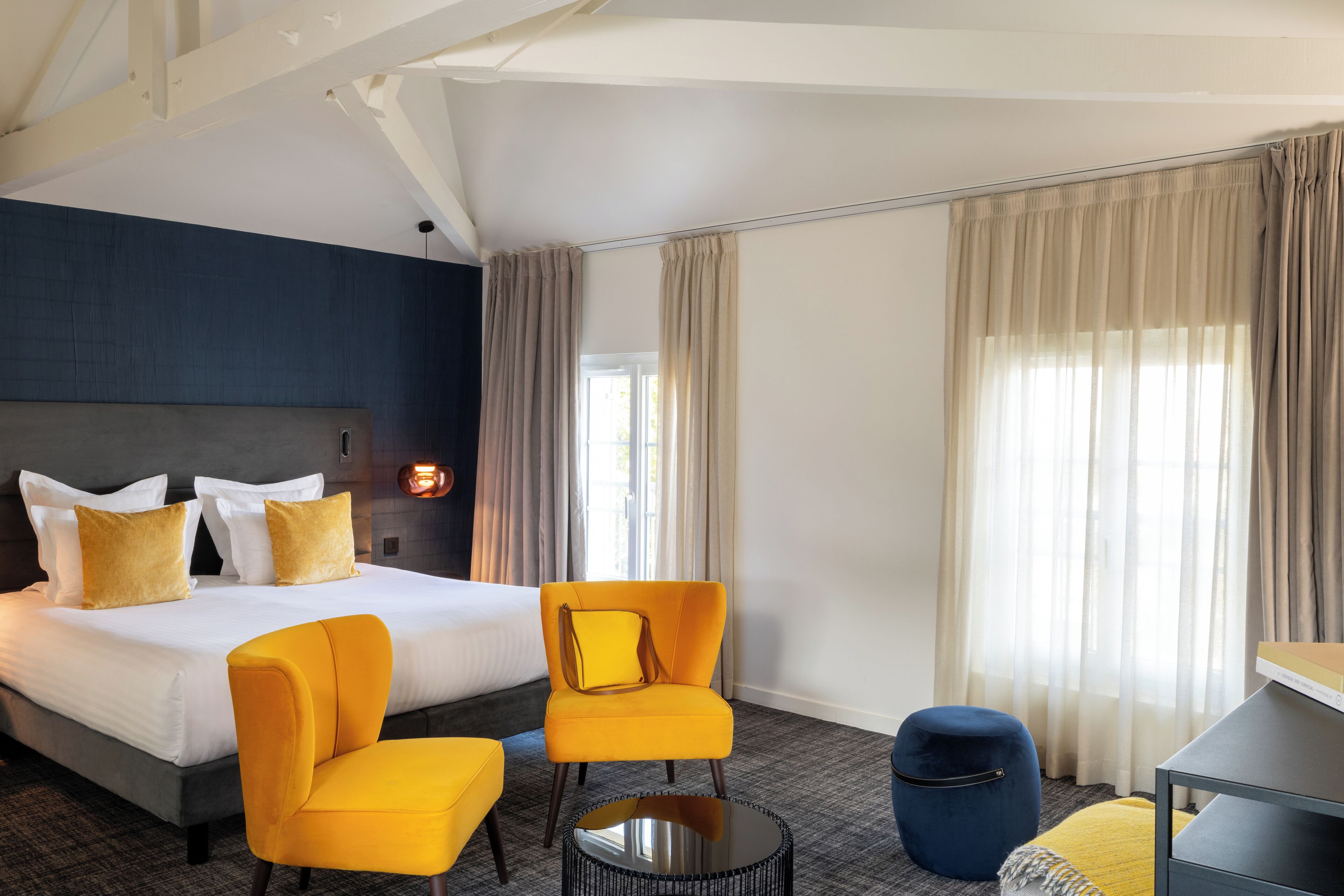 Una habitación de hotel con una cama grande, dos sillones amarillos, una mesa redonda pequeña, un reposapiés azul y un escritorio con una silla.