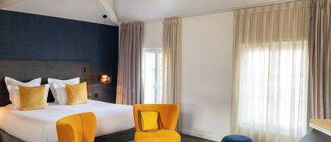 Una habitación de hotel con una cama grande, dos sillones amarillos, una mesa redonda pequeña, un reposapiés azul y un escritorio con una silla.