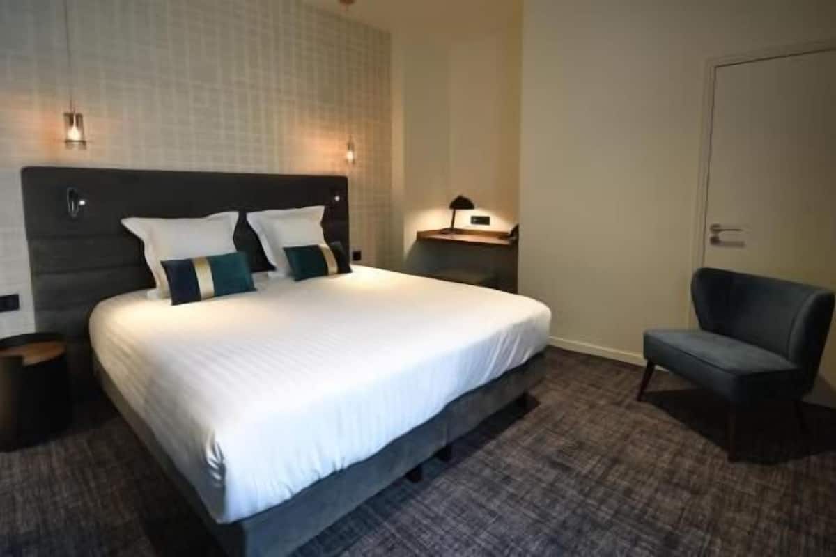 A hotel room with a large bed, a chair, a nightstand, and a wall-mounted lamp.