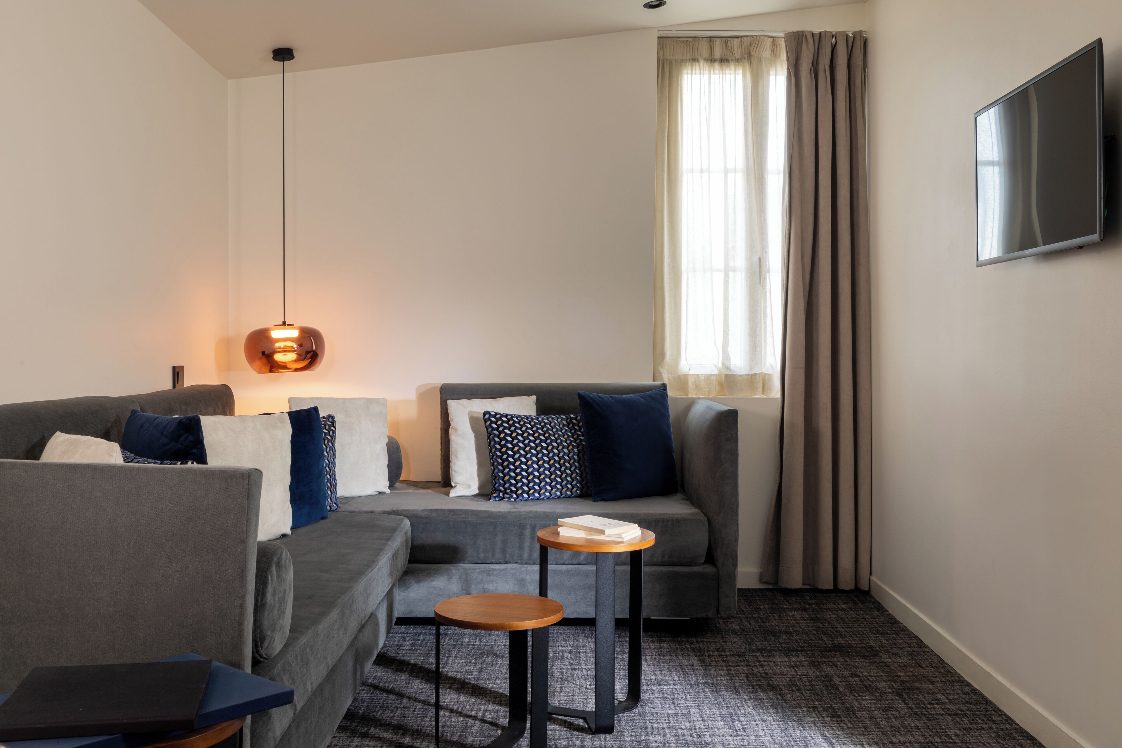 A modern living room with a grey sofa, a small round table, a hanging light fixture, and a TV mounted on the wall.