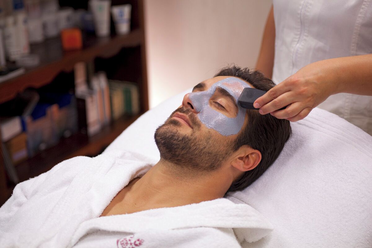 Una persona recostada en una cama con una toalla blanca, un antifaz facial aplicado y un estante con varios objetos al fondo.