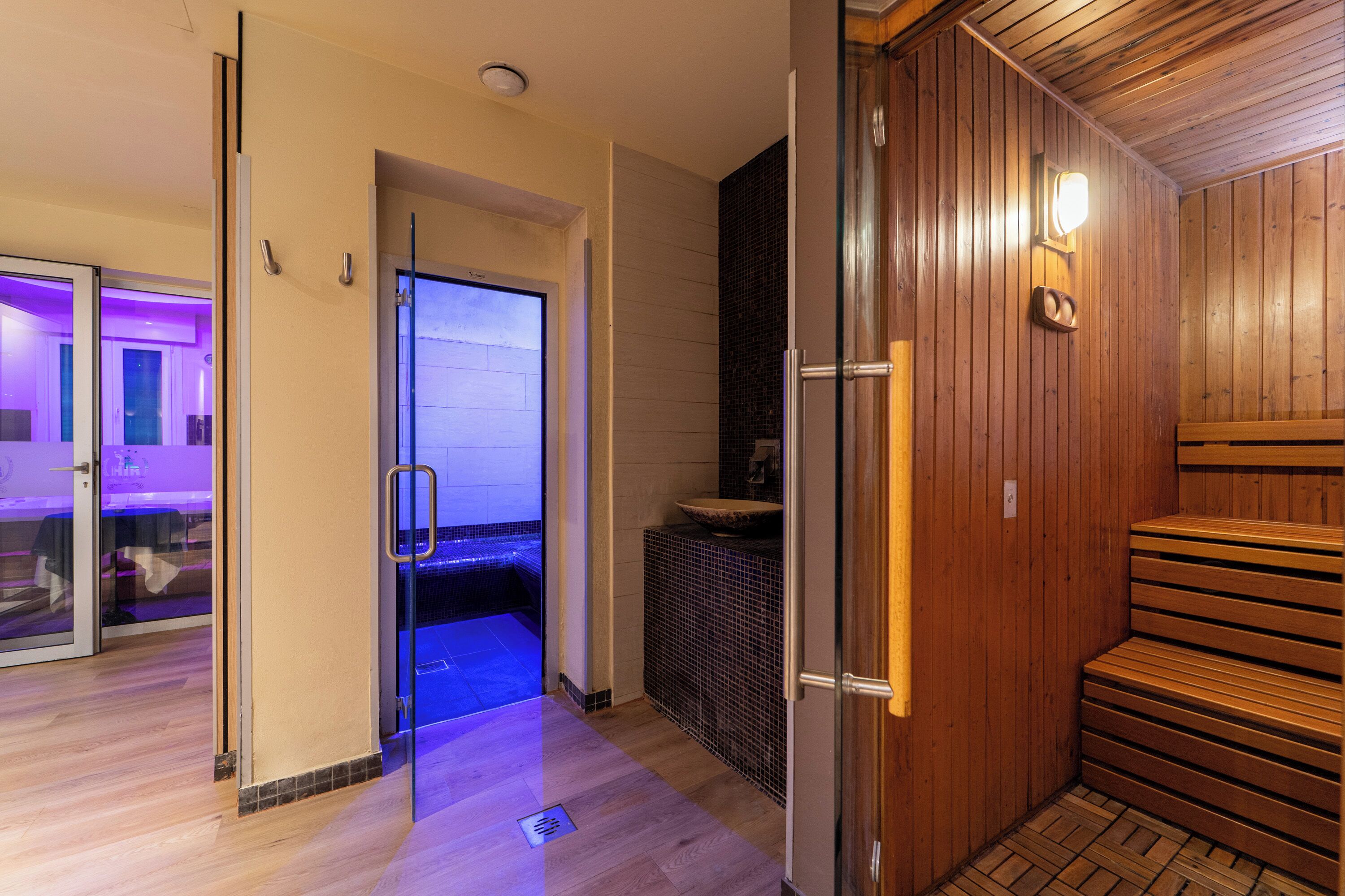 A sauna area with wooden benches, a glass door leading to a room with purple lighting, and a tiled wall.