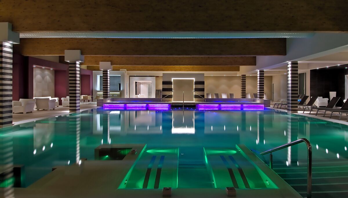 Una piscina interior moderna con un pasillo central, sillas de descanso y una zona de recepción.