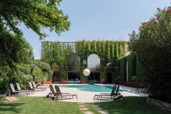 Un área de piscina con sillones de descanso, una piscina cristalina y un jardín con plantas trepadoras y árboles.