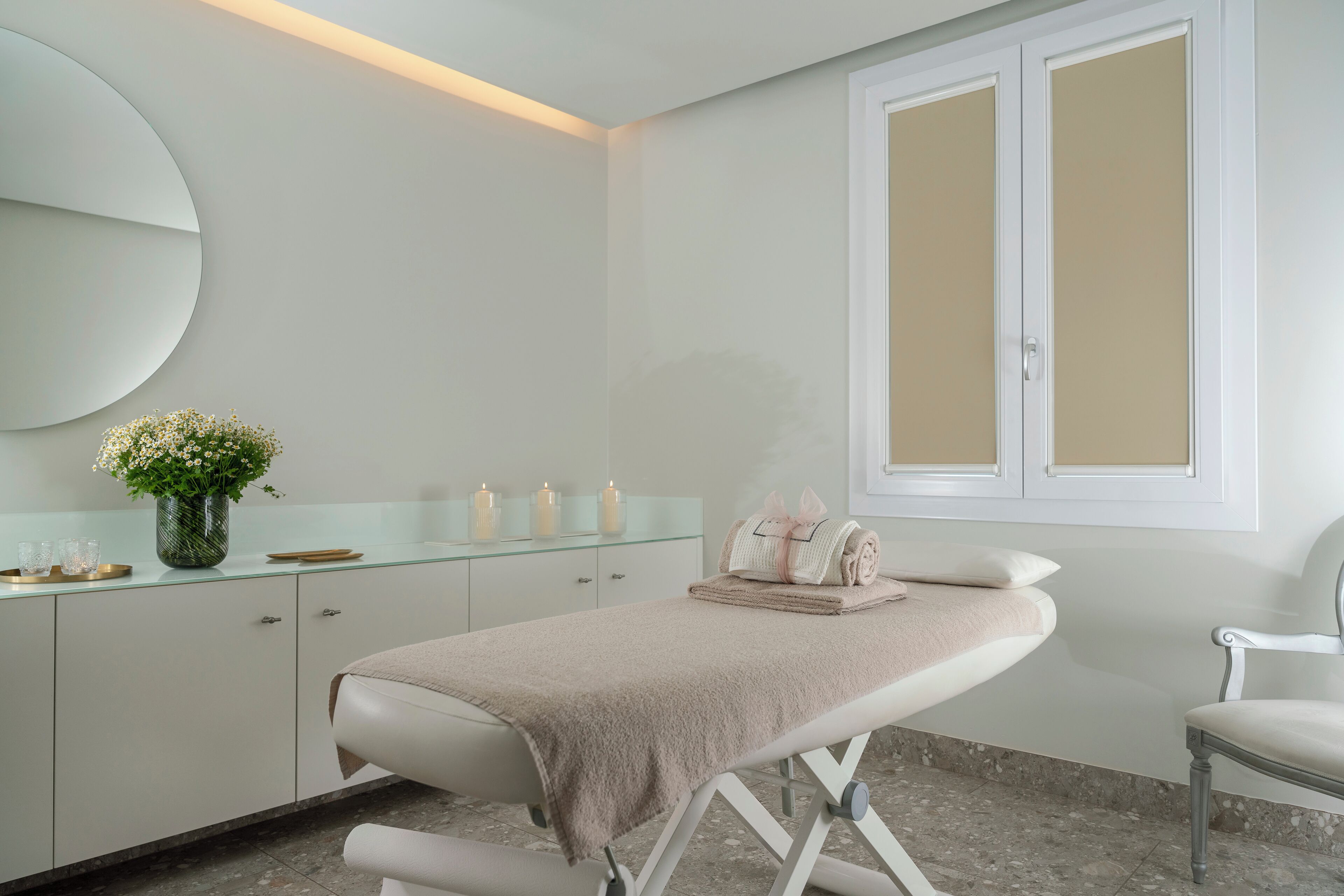 A clean, modern spa room with a massage table, a glass cabinet with candles, and a window with frosted glass panels.