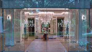 Un interior moderno con una araña, una mesa con flores y el nombre 'MONI PEZZATO' en las puertas de cristal.