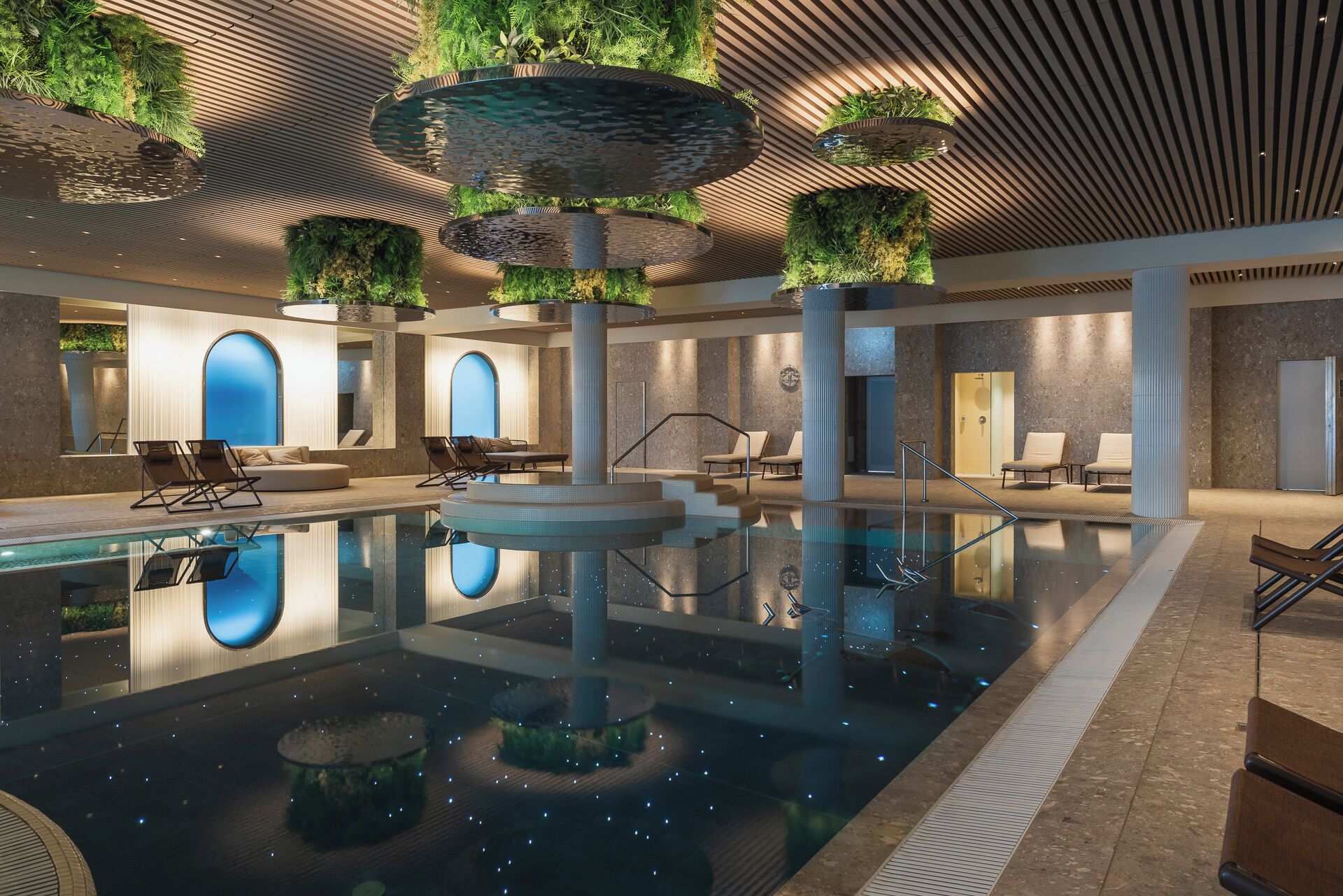 An indoor swimming pool with a central fountain, lounge areas with seating, and a modern architectural design with wooden slats on the ceiling and green plants.