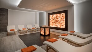 Une salle de spa moderne avec des chaises blanches, un panneau de pierre chauffant et une grande fenêtre ornée d’un motif en briques.