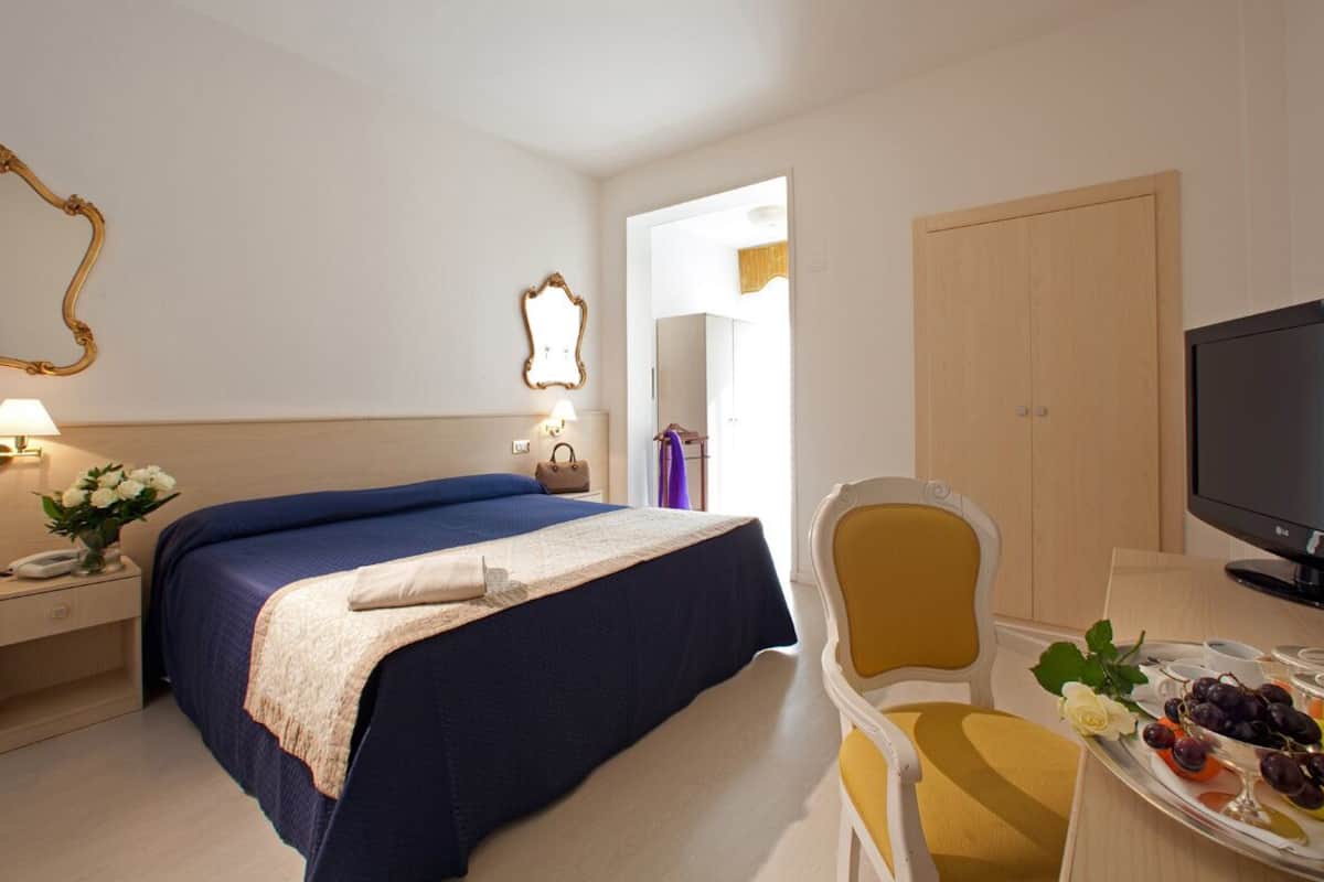 Une chambre d’hôtel équipée d’un lit, d’une table de chevet, d’une lampe, d’un miroir, d’une armoire, d’une télévision et d’un plateau avec des fruits.