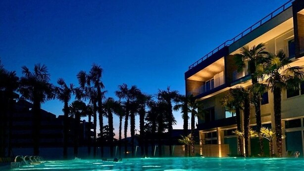 Une piscine à l’eau claire, entourée de palmiers et bordée d’un immeuble à plusieurs étages doté de balcons.