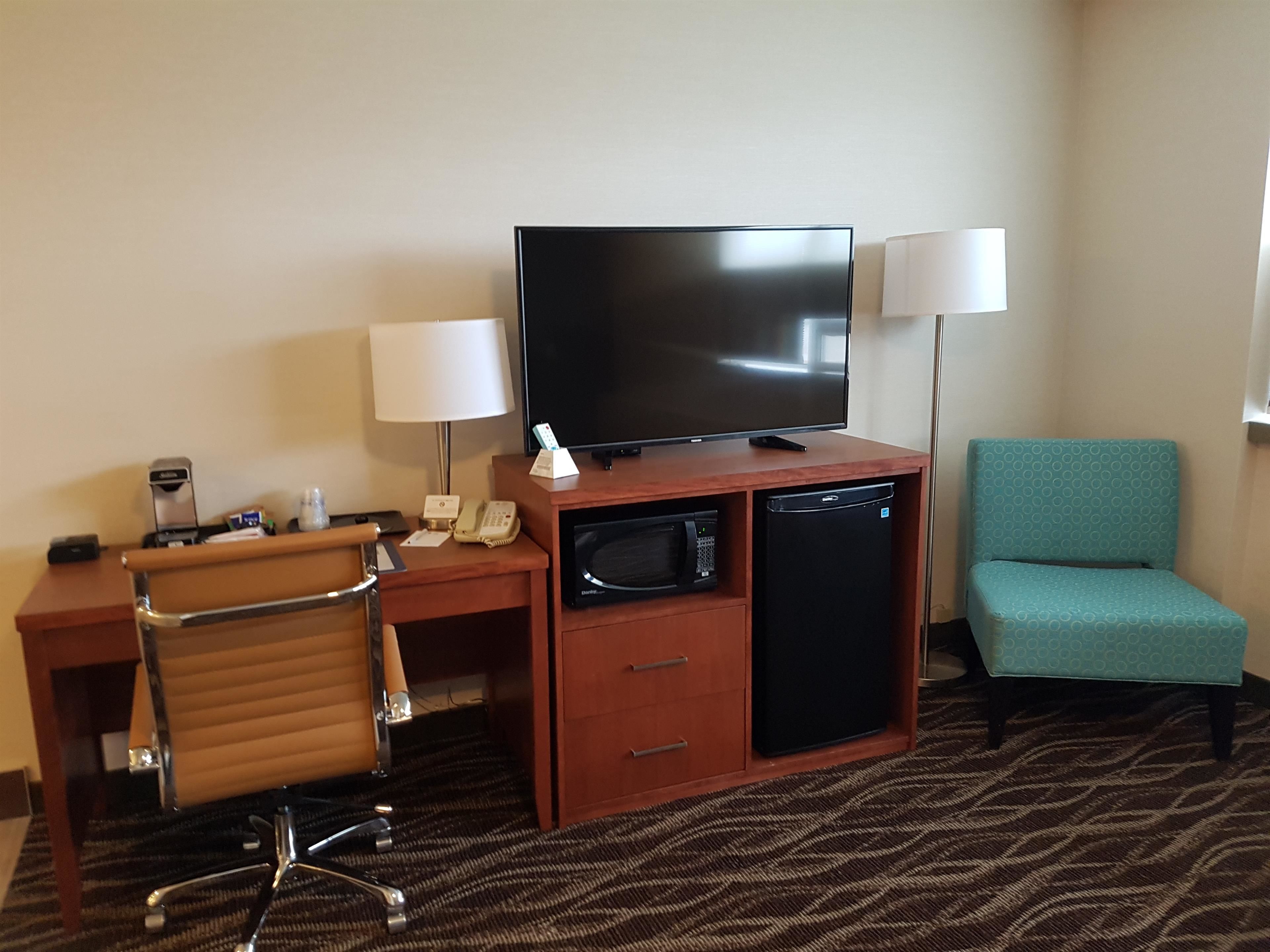 A hotel room with a desk, a television, a microwave, a refrigerator, a chair, and a lamp.