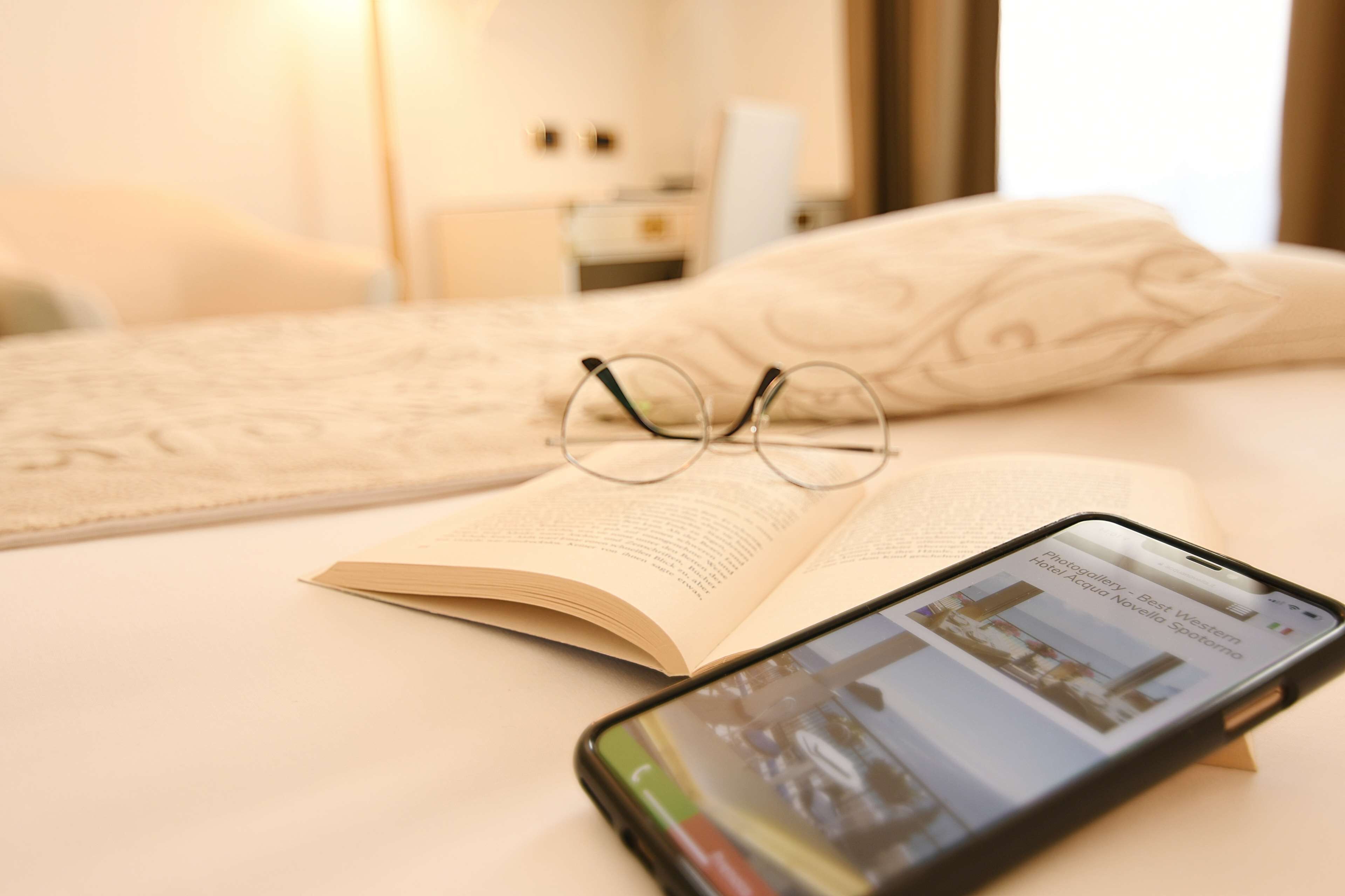 Un letto con un libro, occhiali e uno smartphone che mostra una foto di un hotel, oltre a un comodino con una lampada.
