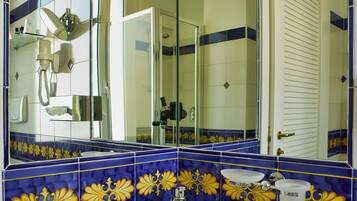 Casa de banho com uma pia branca, azulejos azuis e amarelos, cabine de duche em vidro e um espelho a refletir o espaço.