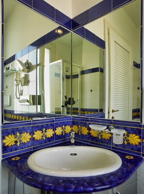 Une salle de bain avec un lavabo blanc, des carreaux bleus et jaunes, une douche en verre et un miroir qui reflète la pièce.