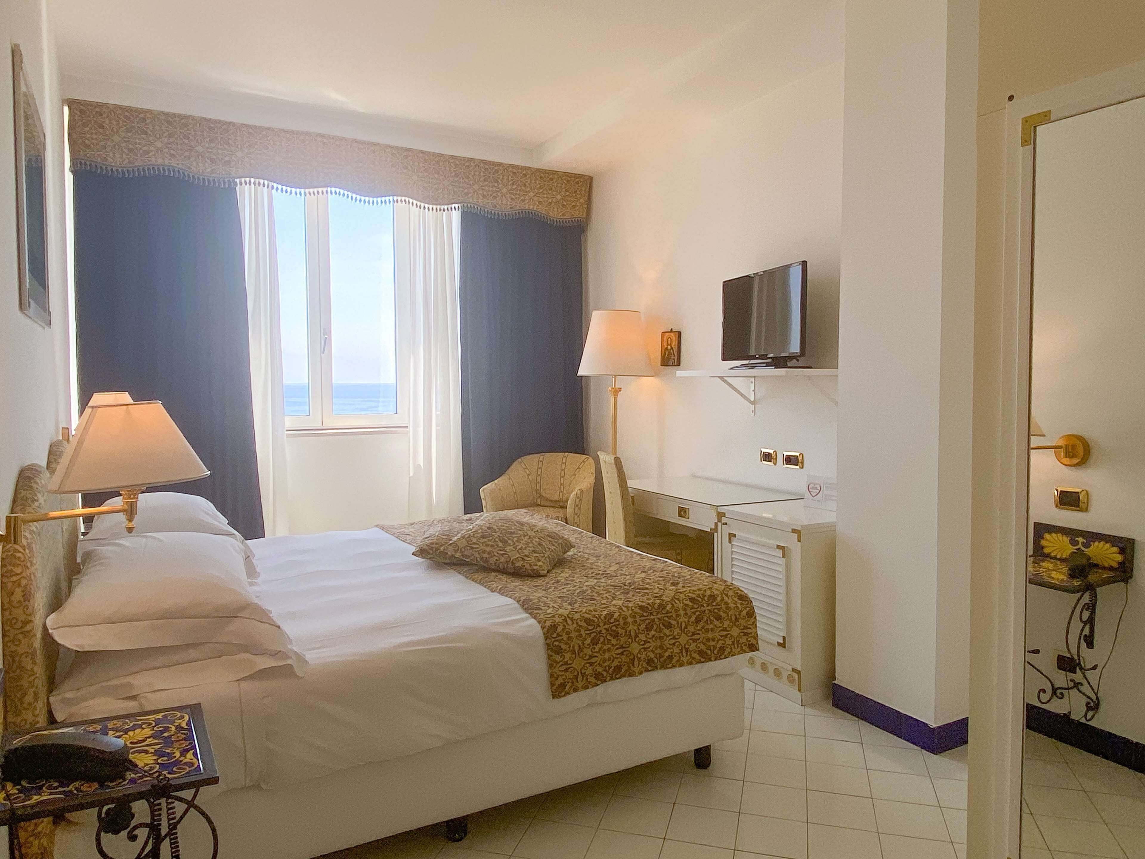 Ein Hotelzimmer mit Bett, Nachttischlampen, einem Schreibtisch mit Fernseher und Meerblick durch das Fenster.