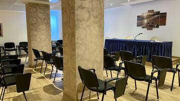 Una sala conferenze con file di sedie, un tavolo blu con un microfono e una parete con un dipinto di un paesaggio.