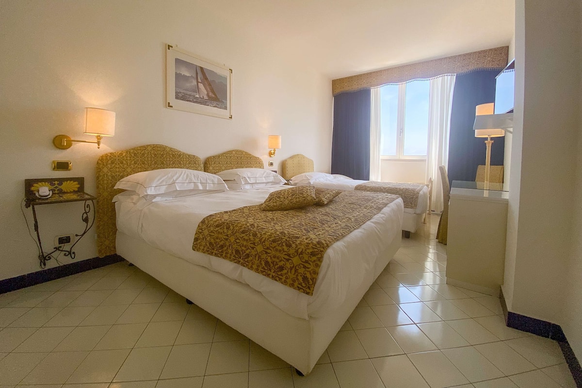 Une chambre d’hôtel avec deux lits, une table de chevet avec une lampe, une petite table avec un téléphone et des objets décoratifs, un tableau au mur et une fenêtre avec des rideaux.