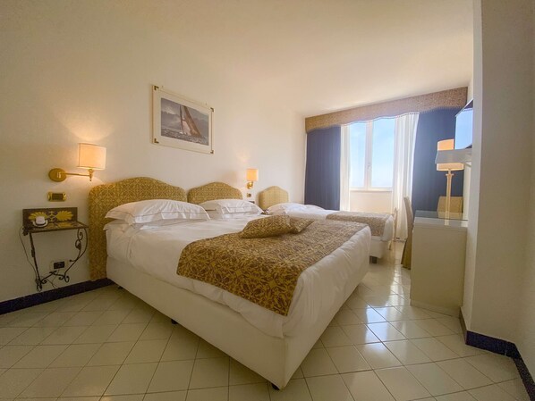 A hotel room with two beds, a bedside table with a lamp, a small table with a phone and decorative items, a painting on the wall, and a window with curtains.
