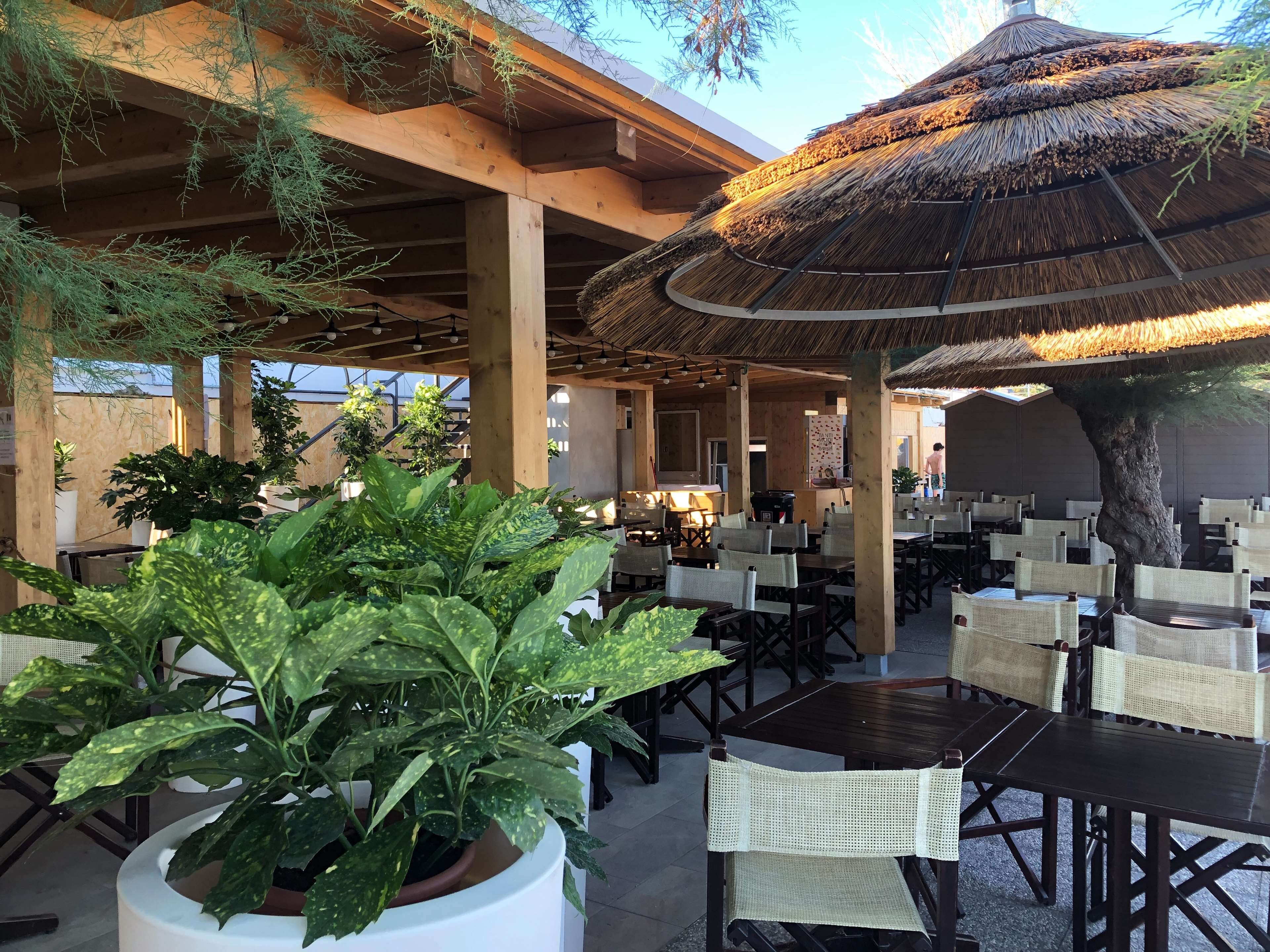 An outdoor seating area with tables and chairs, a thatched roof, and potted plants.