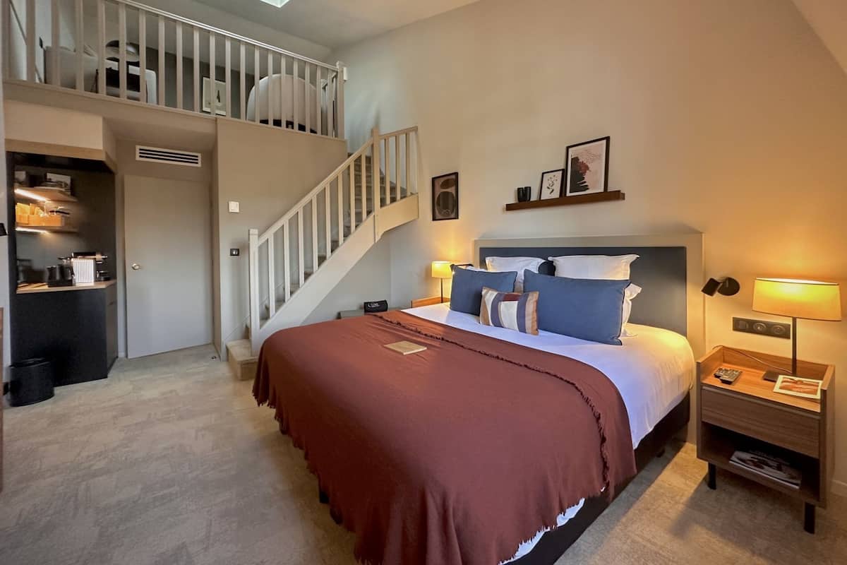 A bedroom with a bed, bedside tables, a nightstand, a staircase, and a small kitchenette.