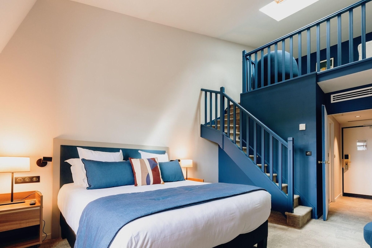 A bedroom with a large bed, a nightstand, a desk, and a blue staircase leading to an upper level.
