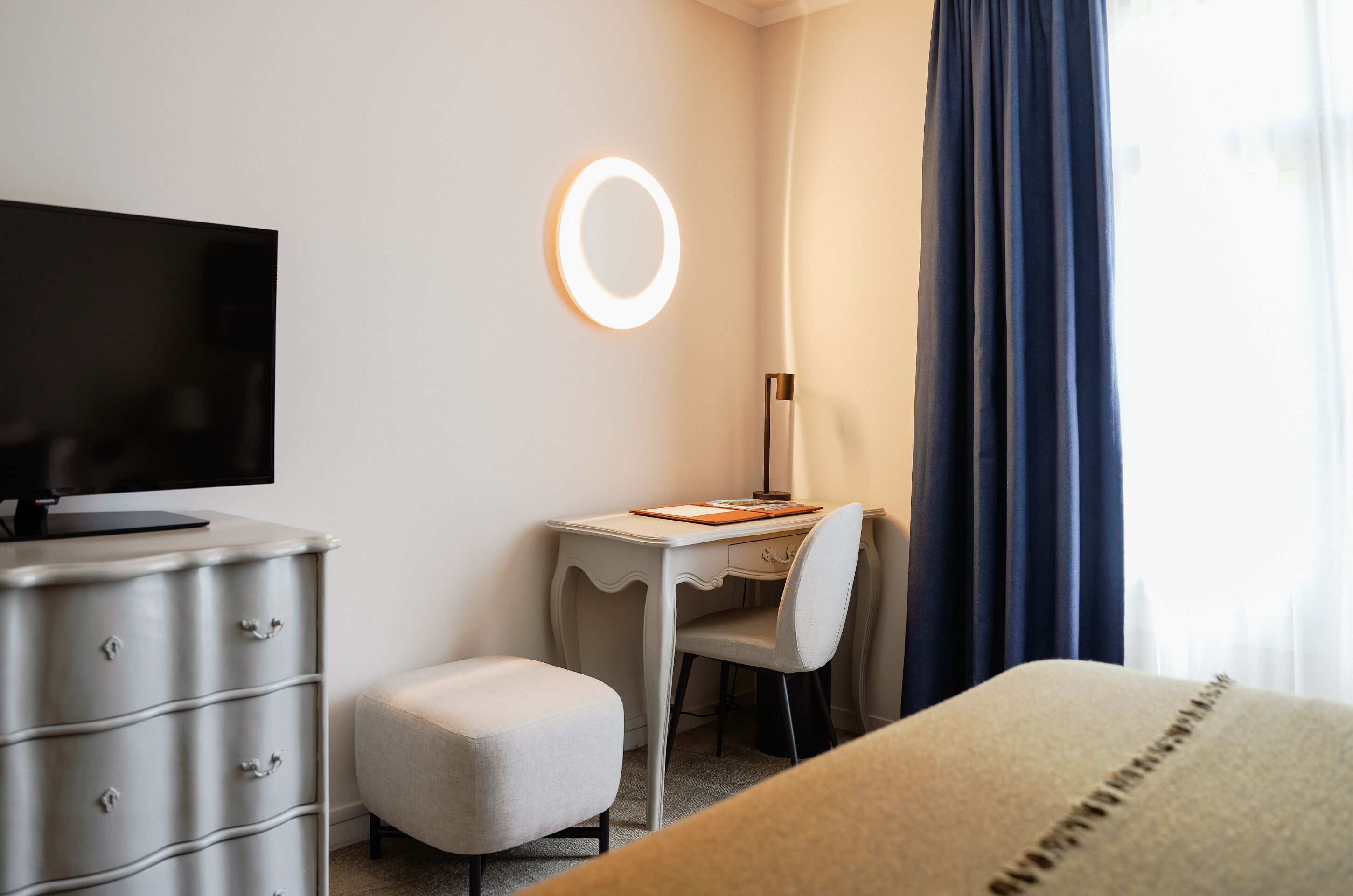 A hotel room with a desk, chair, television, and a bedside table.