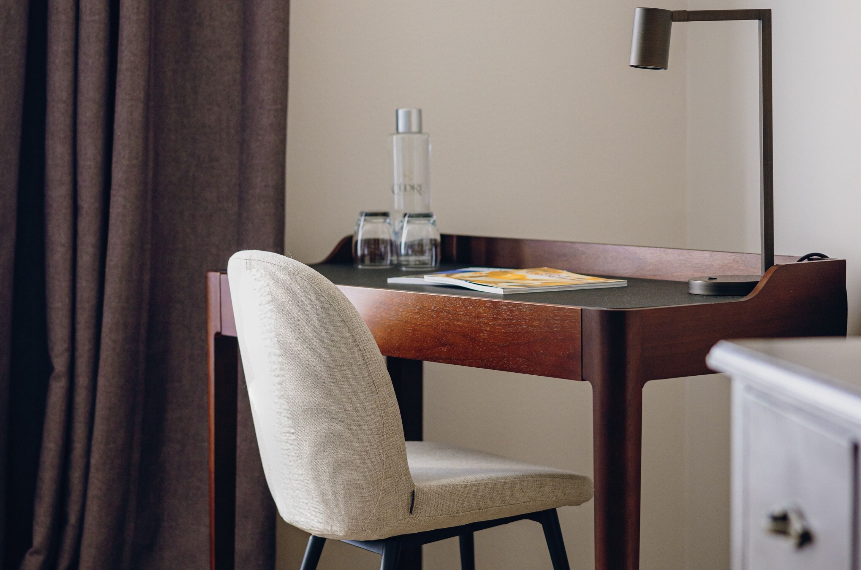 A hotel room with a wooden desk, a chair, a lamp, a bottle, and a book.