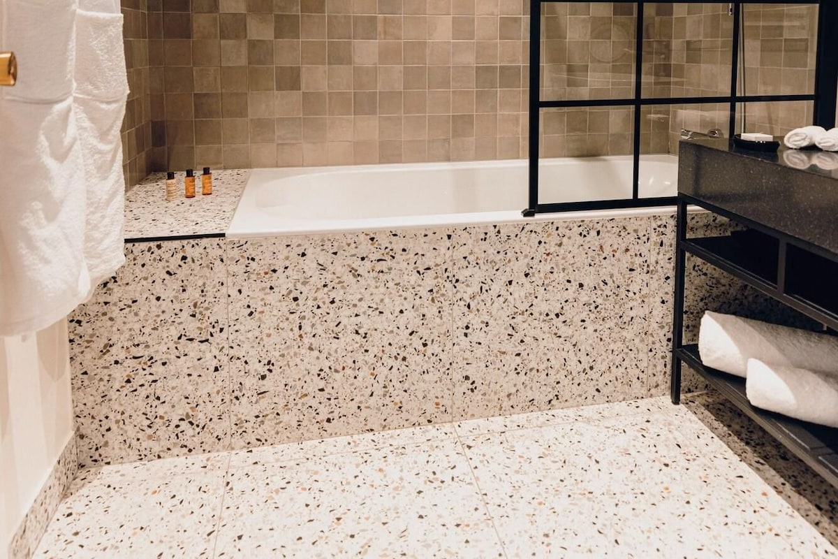 A modern bathroom with a freestanding bathtub, a black metal shower enclosure, and a speckled floor.