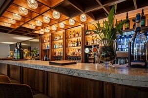 Una zona de bar con estantes de madera llenos de botellas, encimera de mármol y lámparas colgantes esféricas.