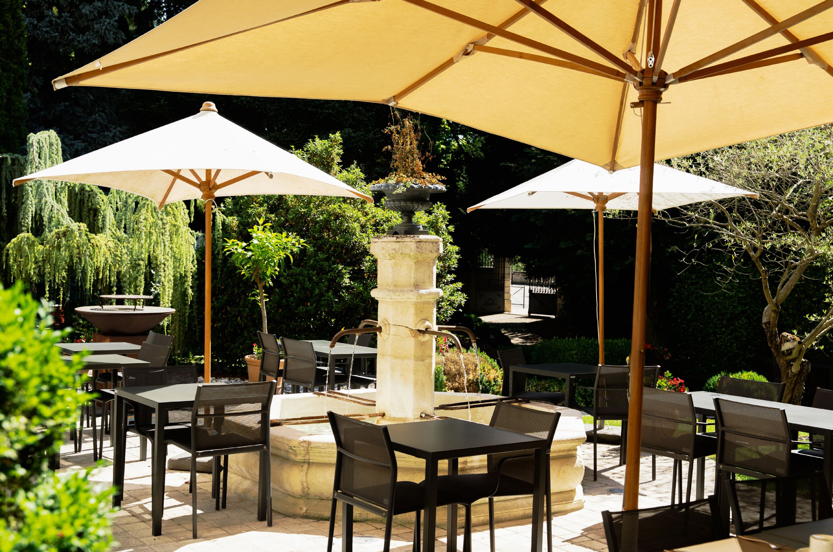 Un patio al aire libre con mesas y sillas, grandes sombrillas y una fuente central.