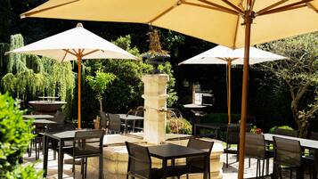 Un patio al aire libre con mesas y sillas, grandes sombrillas y una fuente central.