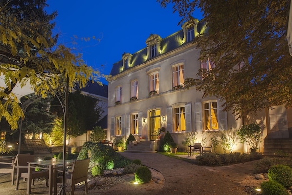 Un bâtiment à deux étages doté d’une entrée bien éclairée, d’une allée pavée et d’un espace extérieur aménagé pour s’asseoir.
