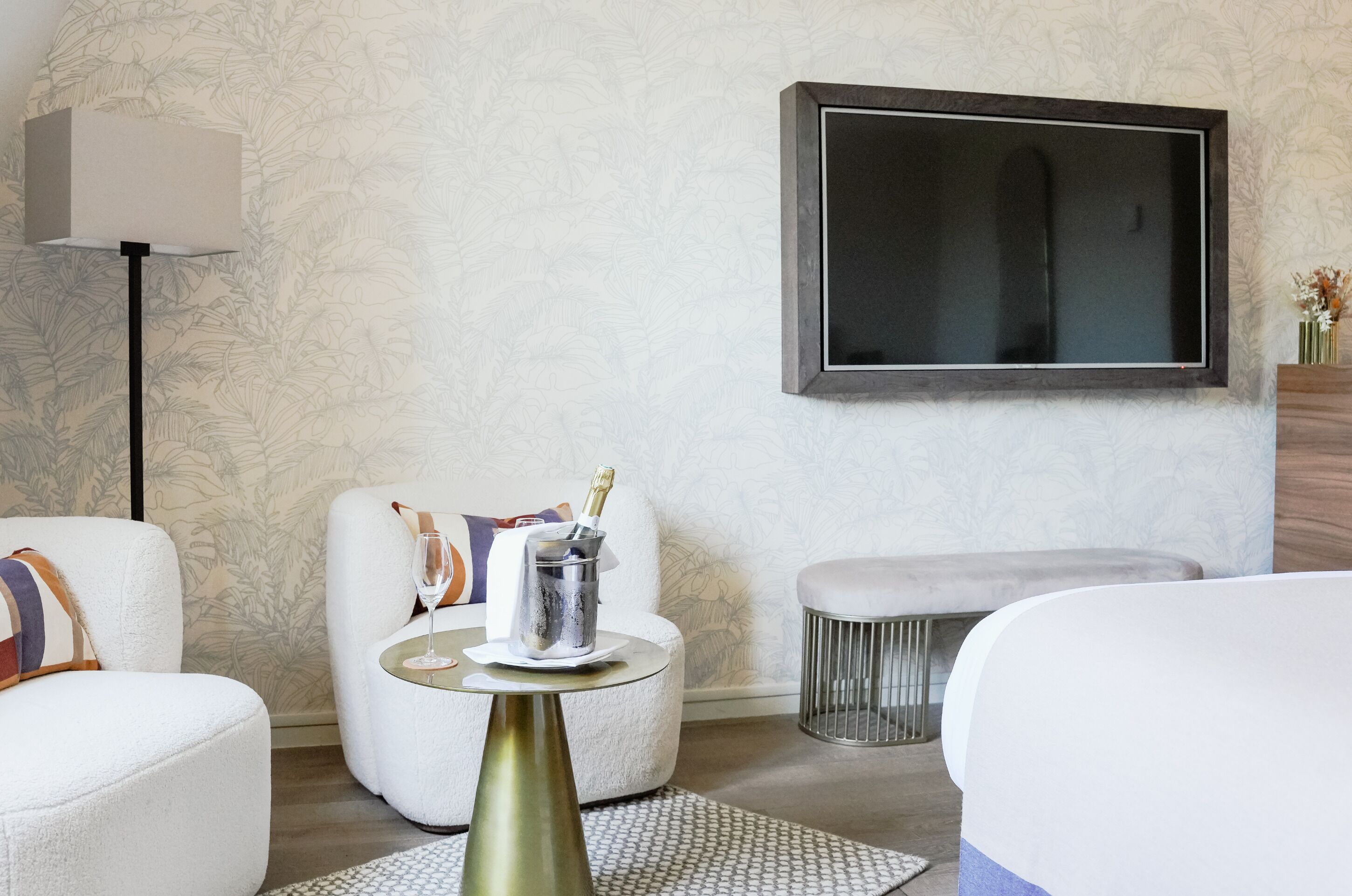 Ein modernes Hotelzimmer mit einem großen Flachbildfernseher, einem runden Beistelltisch mit einer Champagnerflasche und einer gemütlichen Sitzecke mit weißen Sofas und einem gemusterten Teppich.