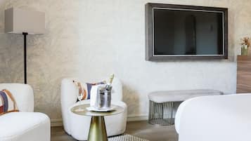Ein modernes Hotelzimmer mit einem großen Flachbildfernseher, einem runden Beistelltisch mit einer Champagnerflasche und einer gemütlichen Sitzecke mit weißen Sofas und einem gemusterten Teppich.