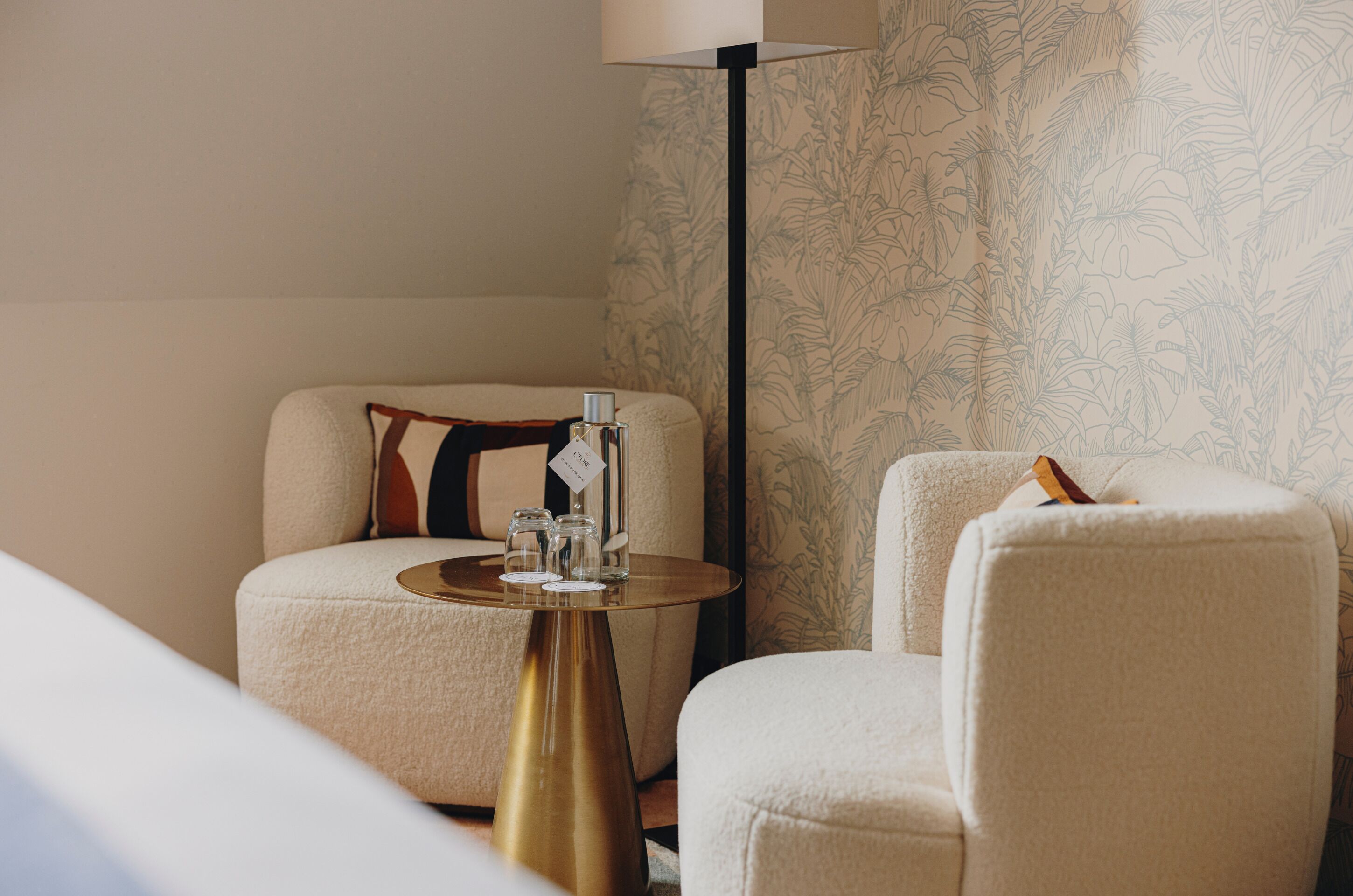A hotel room with a sofa, a small round table with a bottle and glasses, and a floor lamp.