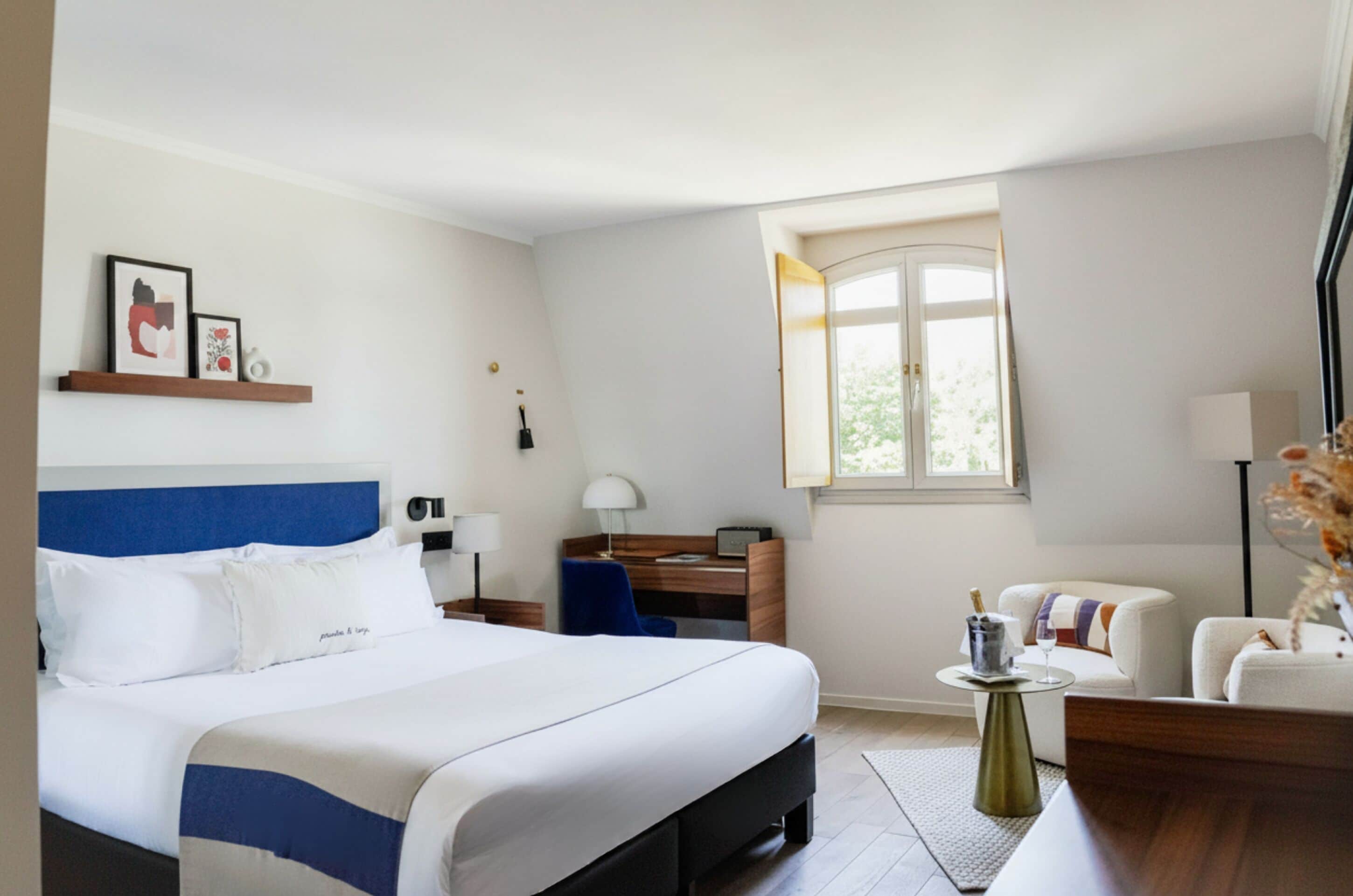 Une chambre d’hôtel avec un lit, un bureau, une chaise, une petite table avec une lampe et une fenêtre.