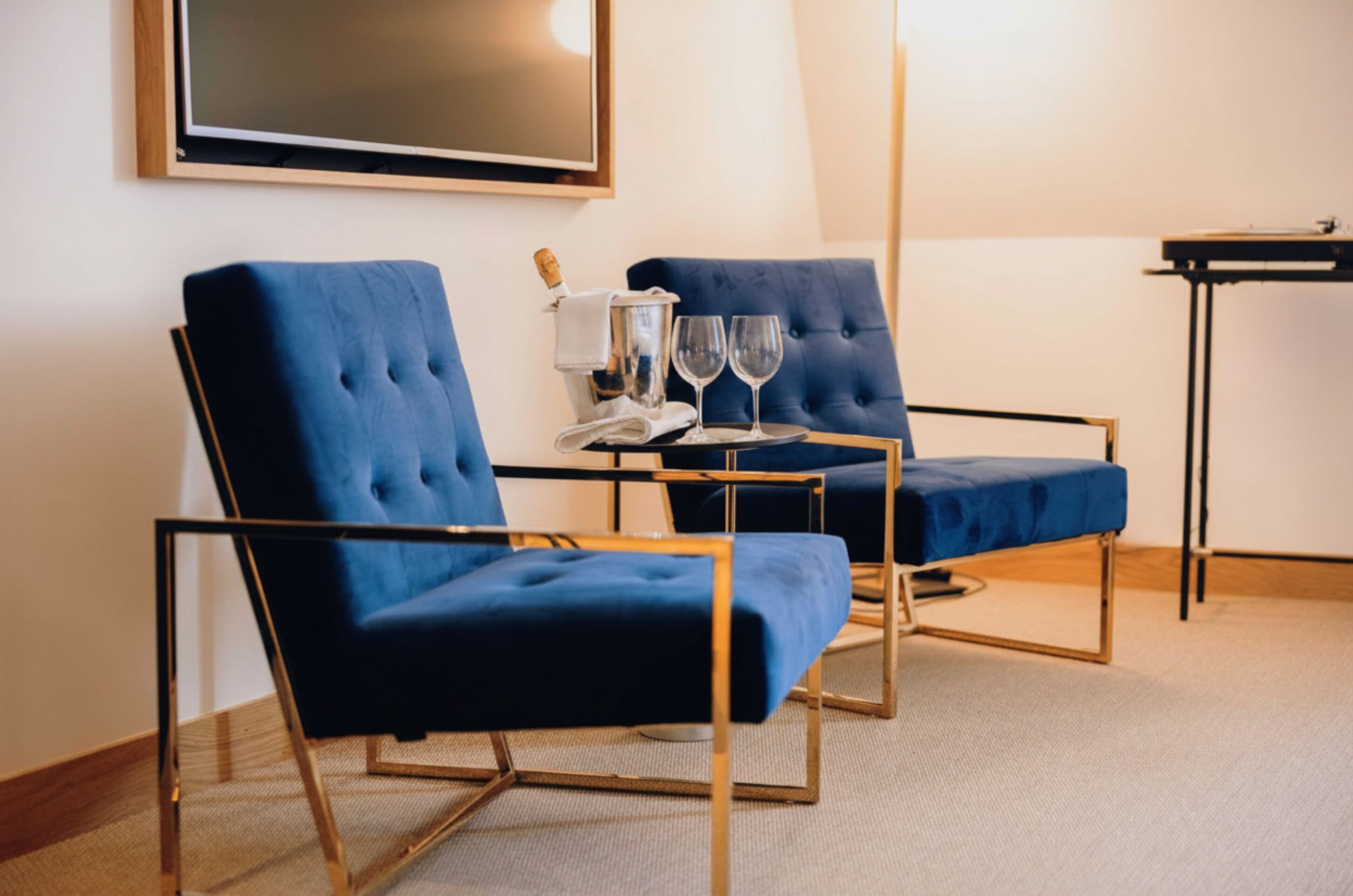 A room with two blue upholstered chairs, a small table with a bottle and glasses, and a large framed picture on the wall.