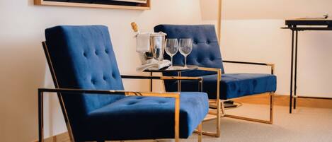 A room with two blue upholstered chairs, a small table with a bottle and glasses, and a large framed picture on the wall.