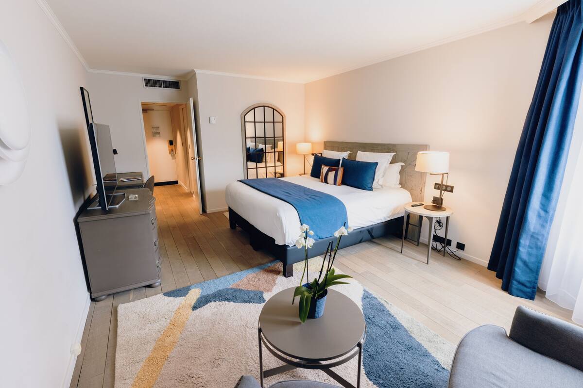 A hotel room with a bed, bedside tables, a desk, a chair, a TV, and a potted plant.