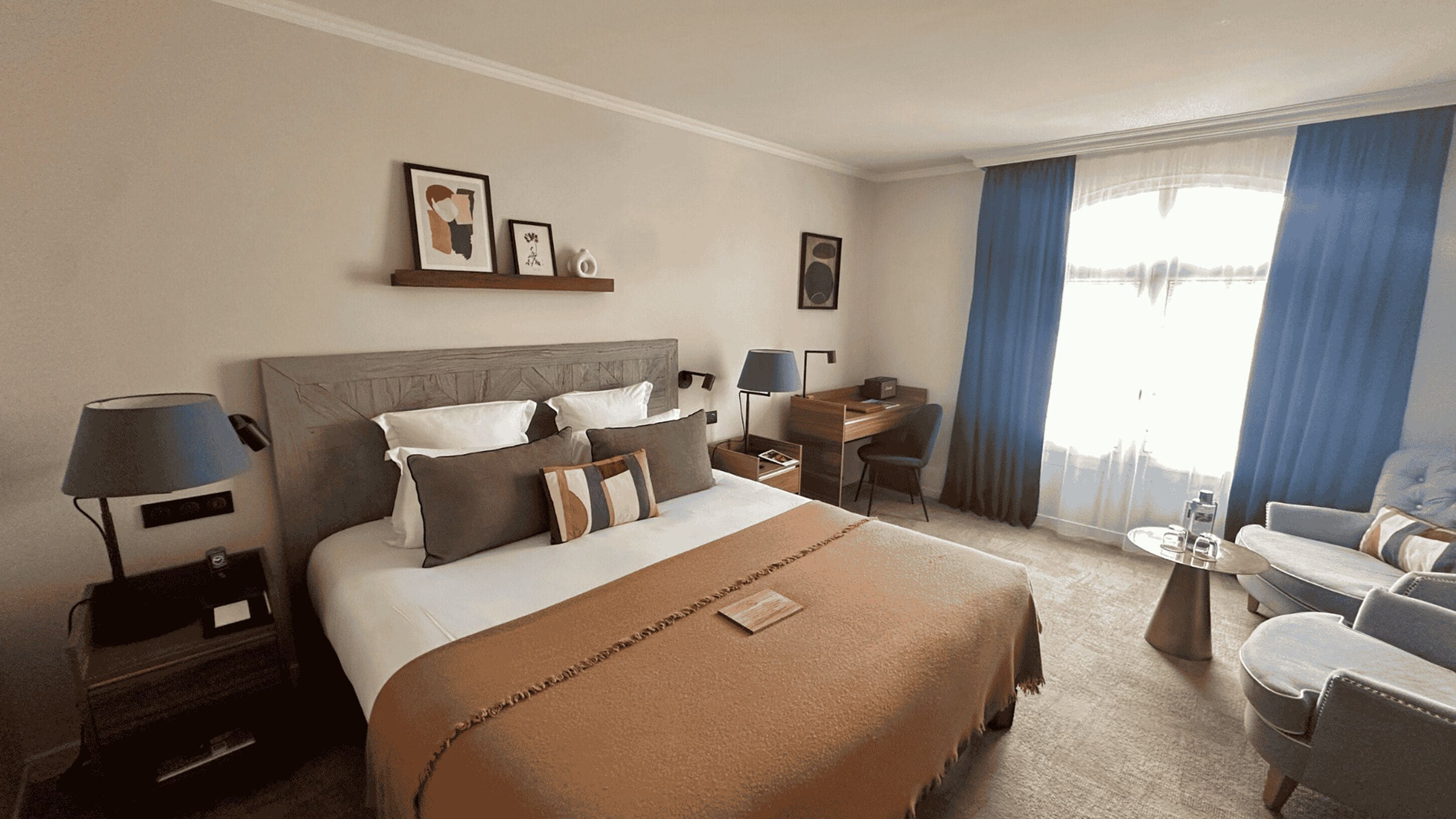 A hotel room with a large bed, a desk, a chair, and a small table. There are two lamps, a picture on the wall, and a window with curtains.