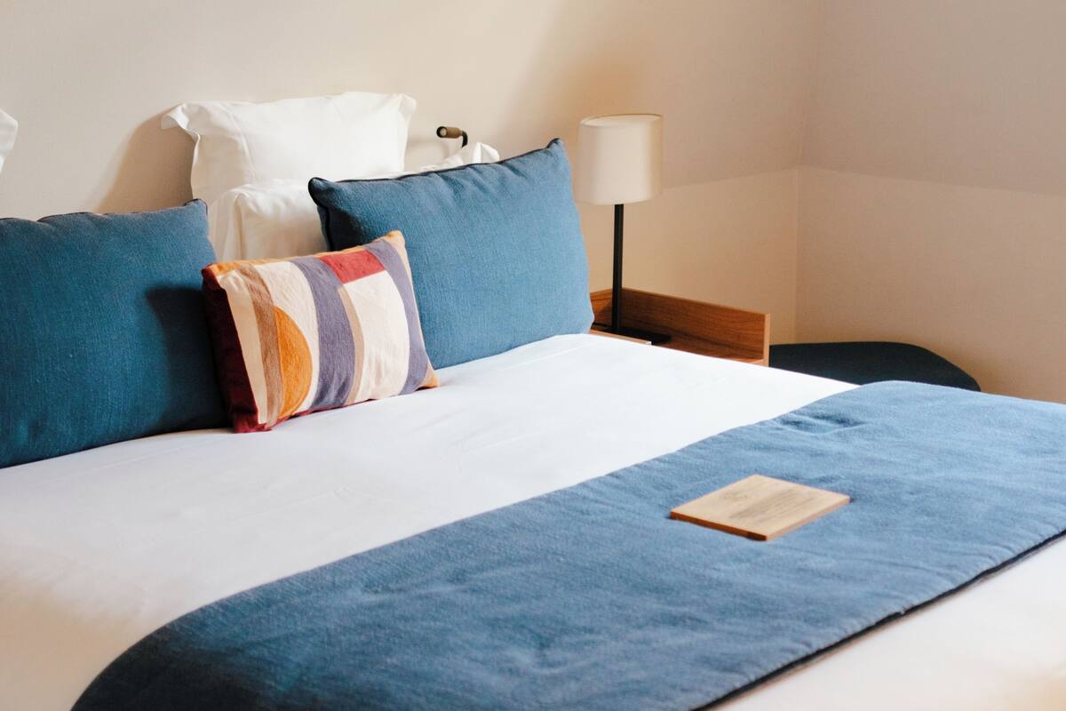 A neatly made bed with blue bedding, a colorful pillow, and a bedside table with a lamp.