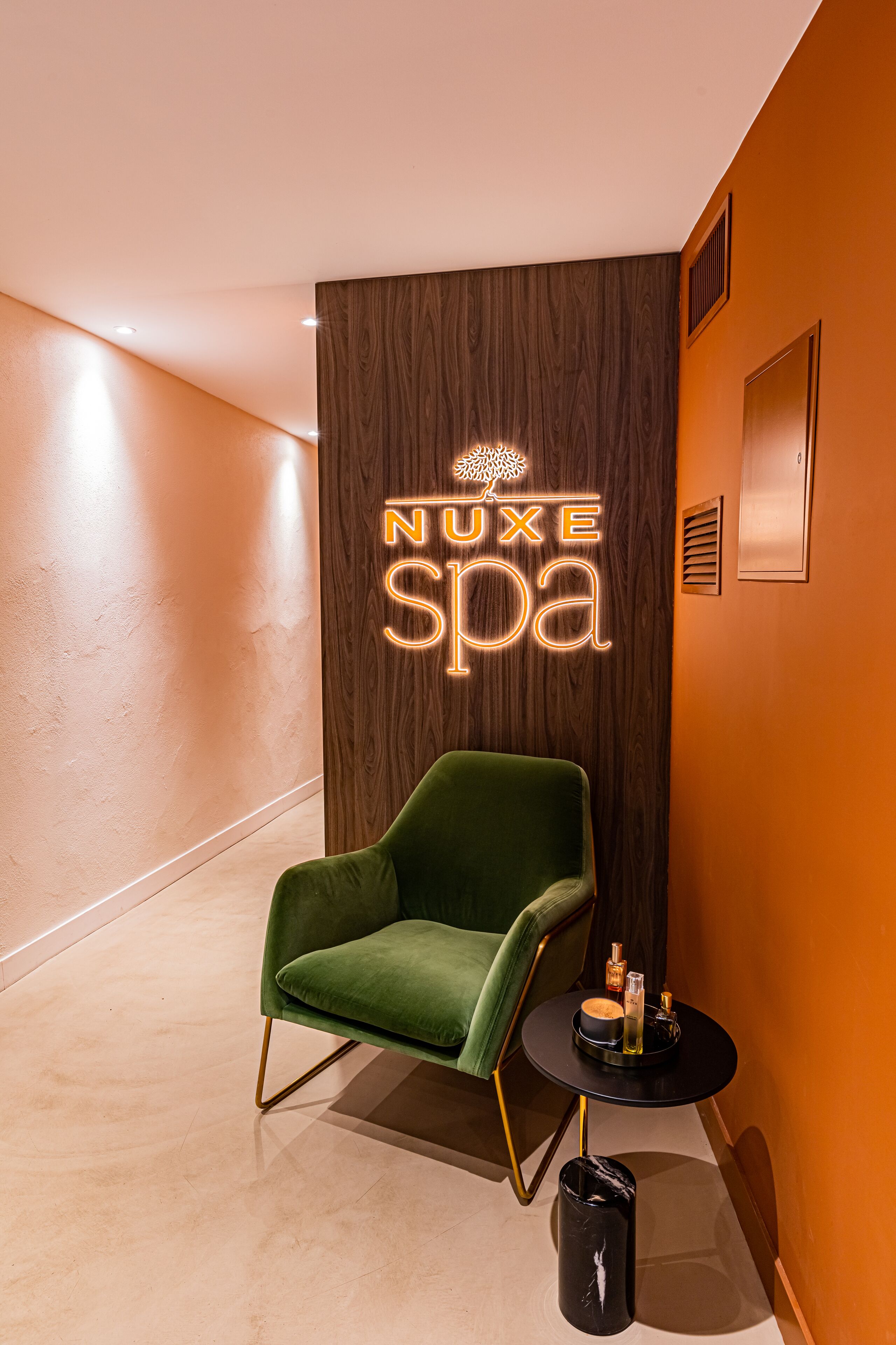 Zona de spa con una silla verde, una mesita con botellas y un letrero de neón que dice "NUXE spa".