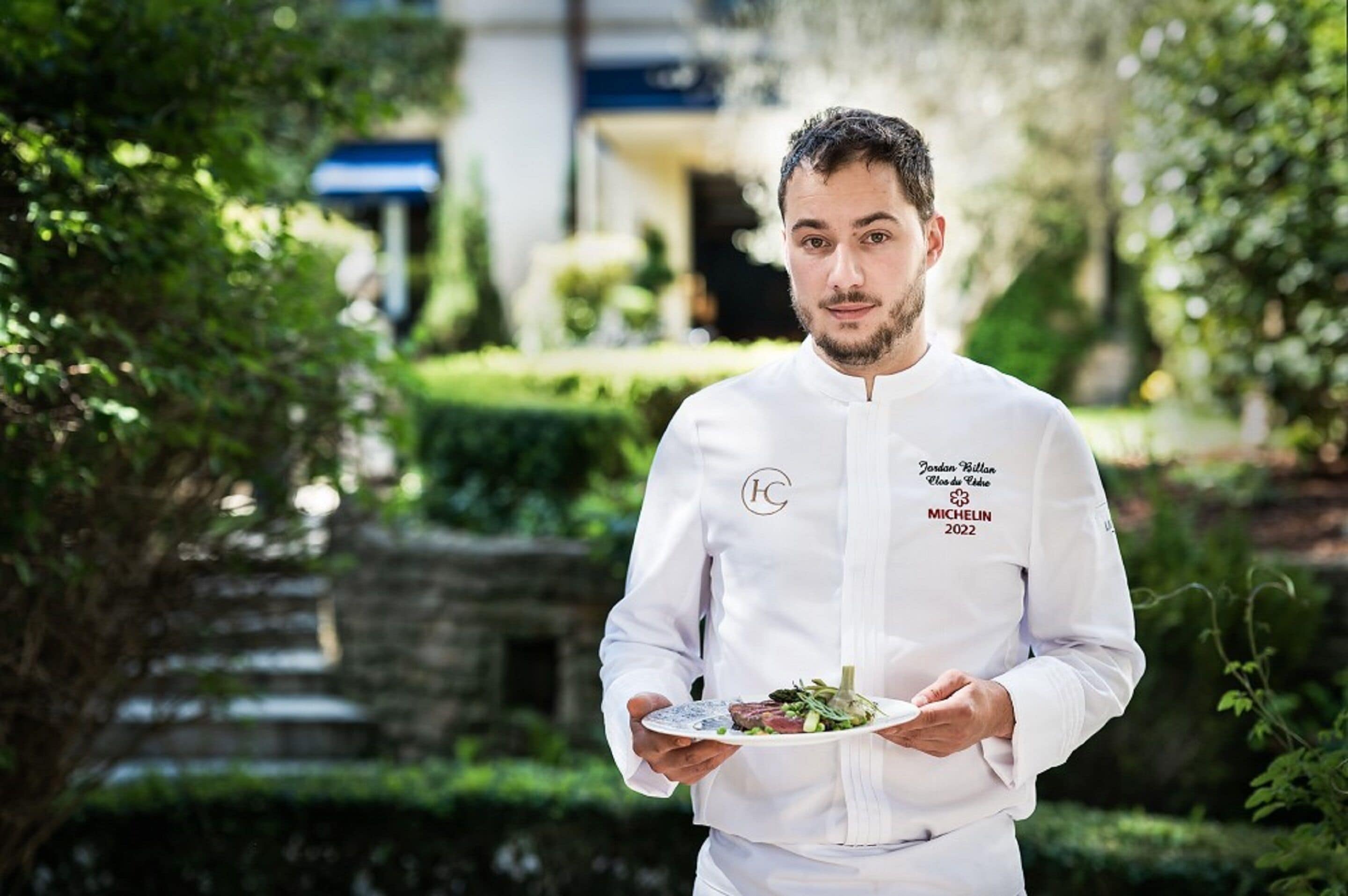 A chef in a white uniform holding a plate of food outdoors with greenery and a building in the background.