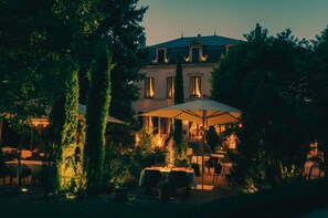 An outdoor dining area with tables and umbrellas, surrounded by trees and a building with large windows. - Cèdre Beaune, A Beauvallon Hotel & Spa (Beaune)