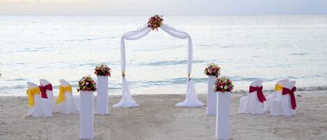 Une arche décorée pour un mariage en plein air, installée sur une plage, avec des chaises blanches et des décorations florales.