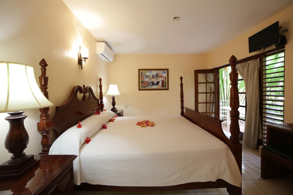 Uma cama bem arrumada com colcha branca e flores decorativas, uma mesa de cabeceira de madeira com abajur, uma televisão presa à parede e uma janela com cortinas.