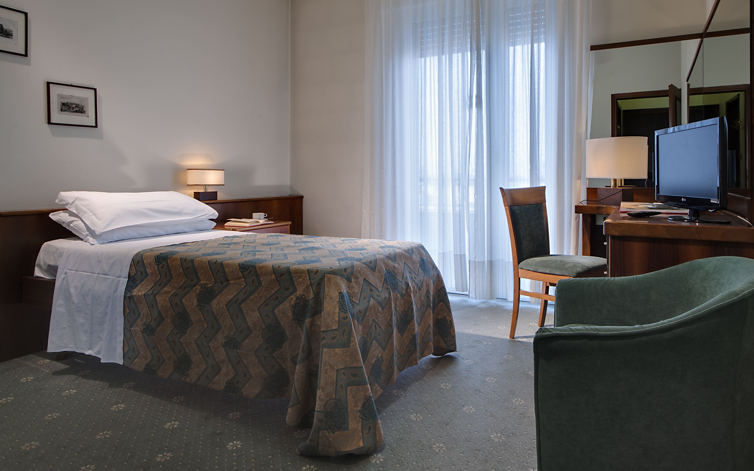 Quarto de hotel com cama, escrivaninha com computador, cadeira e janela com cortinas.