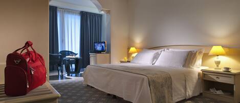 A hotel room with a bed, a desk with a computer, a chair, a red suitcase, and a TV on the desk.